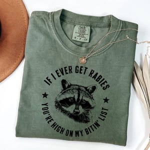 May include: An olive green t-shirt with the text "IF I EVER GET RABIES YOU'RE HIGH ON MY BITIN' LIST" surrounding a raccoon illustration. The shirt has a crew neck and short sleeves. A gold necklace is draped across the shirt.