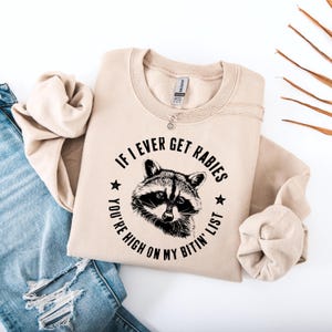 May include: Beige sweatshirt with the text "IF I EVER GET RABIES YOU'RE HIGH ON MY BITIN' LIST" surrounding a raccoon illustration. The sweatshirt is folded, with a silver necklace visible. A pair of blue jeans is partially visible in the background.