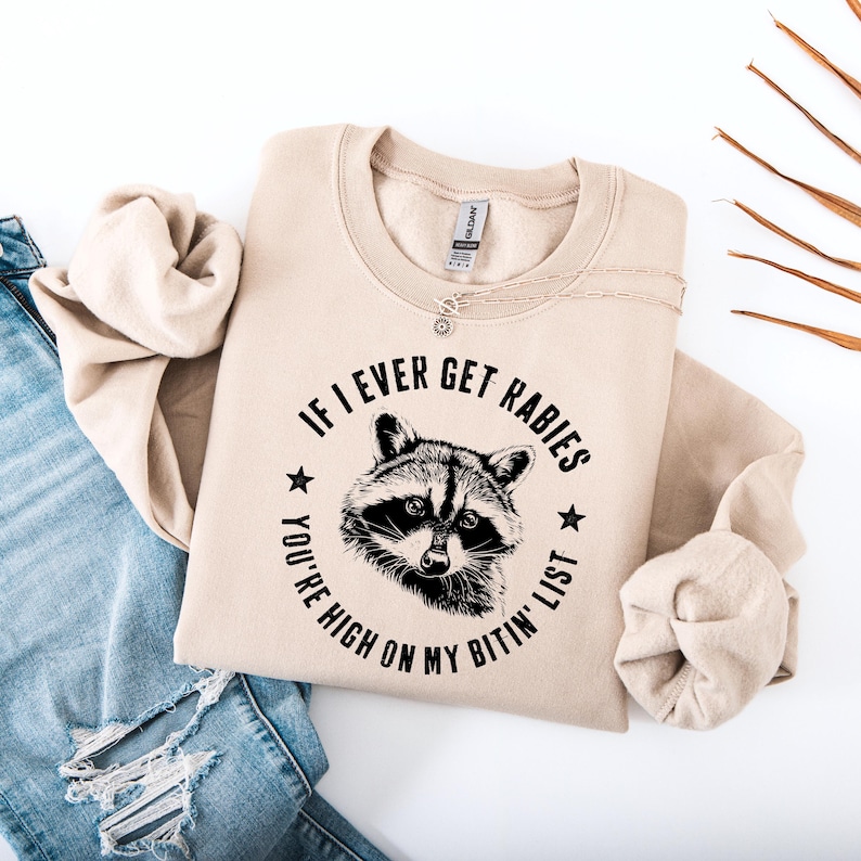 May include: Beige sweatshirt with the text "IF I EVER GET RABIES YOU'RE HIGH ON MY BITIN' LIST" surrounding a raccoon illustration. The sweatshirt is folded, with a silver necklace visible. A pair of blue jeans is partially visible in the background.