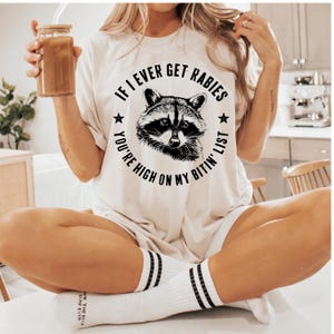 May include: Cream-colored t-shirt featuring a raccoon illustration with the text "If I Ever Get Rabies, You're High On My Bitin' List." The shirt is paired with white socks with black stripes. A glass jar with a drink is held in the hand.