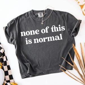 May include: Dark gray t-shirt with the phrase "none of this is normal" in white block letters. The shirt has rolled-up sleeves and a crew neck. A silver necklace with a pendant is draped on the shirt.