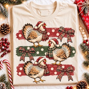 Peut inclure: Un t-shirt couleur crème avec une impression graphique de trois poules portant des bonnets de Père Noël rouges et des écharpes à carreaux. Le motif comprend des bandes horizontales brossées rouges et vertes à pois blancs et trois nœuds.