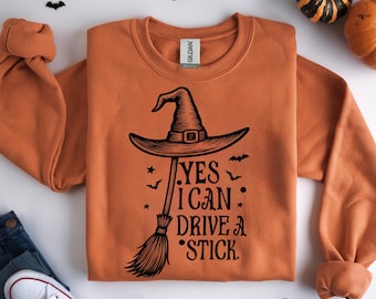 May include: Orange sweatshirt with a black witch hat and broom graphic. The text reads "YES I CAN DRIVE A STICK." The sweatshirt is folded, with a pair of jeans and a white sneaker visible in the lower left corner. Decorative pumpkins and bats are scattered around.