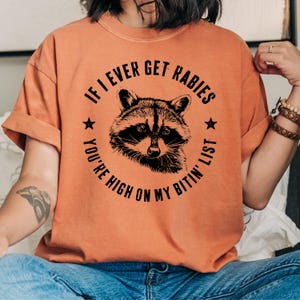 May include: Orange t-shirt featuring a raccoon illustration within a circular design. The text reads "IF I EVER GET RABIES YOU'RE HIGH ON MY BITIN' LIST". The shirt has short sleeves and a relaxed fit.