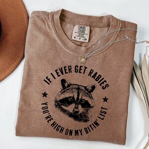 May include: A brown Comfort Colors t-shirt with the text "IF I EVER GET RABIES YOU'RE HIGH ON MY BITIN' LIST" surrounding a raccoon graphic. A silver necklace with a small pendant is draped across the shirt.