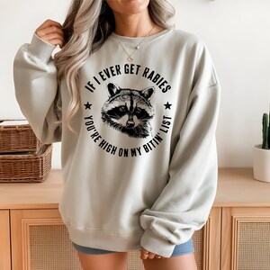 May include: A light gray sweatshirt with a raccoon graphic and the text "IF I EVER GET RABIES YOU'RE HIGH ON MY BITIN' LIST". The raccoon is in a circular design with stars.