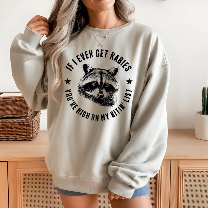 May include: A light gray sweatshirt with a raccoon graphic and the text "IF I EVER GET RABIES YOU'RE HIGH ON MY BITIN' LIST". The raccoon is in a circular design with stars.
