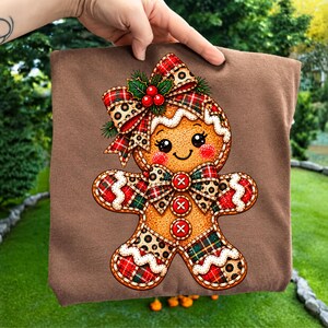 Könnte beinhalten: Ein braunes T-Shirt mit einer Grafik eines Lebkuchenmanns, der mit einer Schleife in Schottenkaro- und Leopardenmuster, einer passenden Fliege und drei roten Knöpfen verziert ist. Die Figur hat weiße Zuckergussakzente, grüne Stechpalmenblätter und rote Beeren.
