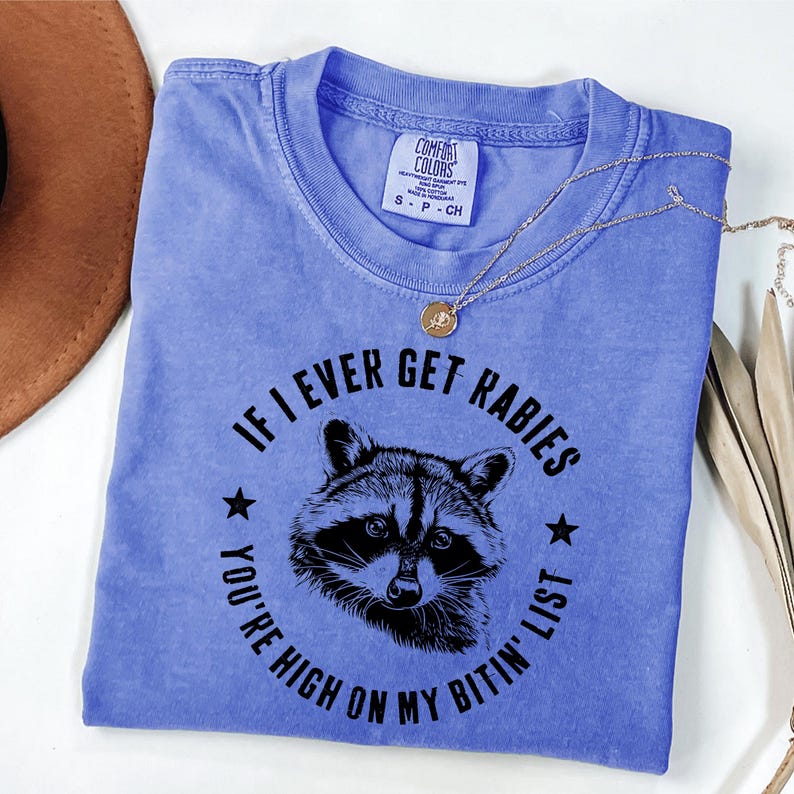 May include: A periwinkle blue t-shirt with the text "IF I EVER GET RABIES YOU'RE HIGH ON MY BITIN' LIST" surrounding a raccoon illustration. The shirt has a crew neck and short sleeves. A gold necklace is draped across the shirt.