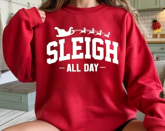 Könnte beinhalten: Ein rotes Sweatshirt mit dem weißen Text "SLEIGH ALL DAY". Das Design beinhaltet einen Schlitten und Rentiere. Das Sweatshirt hat einen Rundhalsausschnitt.