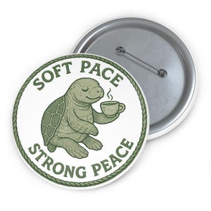 May include: A round button with a green turtle illustration holding a cup of coffee. The button has the text "SOFT PACE STRONG PEACE" in green. The button is white with a silver pin back.