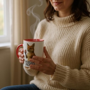 Op de afbeelding: Witte keramische mok met een rode binnenkant en oor. De mok heeft een illustratie van een vos die een appel vasthoudt, met de tekst "FEELIN' FOXY" eronder. Stoom stijgt op uit de mok, wat duidt op een warme drank.