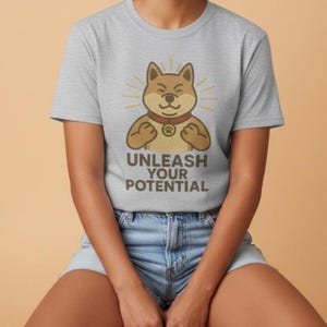 Puede incluir: Camiseta gris claro con un perro Shiba Inu de dibujos animados con un collar rojo y una huella de pata. El perro flexiona sus brazos con el texto "UNLEASH YOUR POTENTIAL" debajo. La camiseta es de cuello redondo.