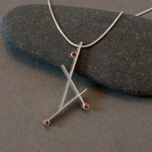 May include: A silver pendant necklace with a geometric design. The pendant features an abstract shape with three small, round, red gemstones. The necklace is displayed on a dark gray stone surface.