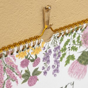 May include: A wall calendar hanging from a gold metal hook, featuring a white page with botanical illustrations of pink clover, purple wisteria, yellow flowers, and green leaves. The calendar is bound with a gold wire spiral coil at the top.