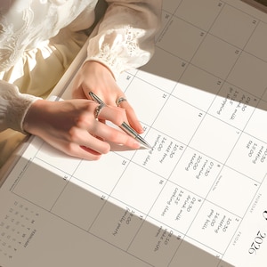 May include: A white paper wall calendar for the year 2026 with black grid lines and printed text. The calendar features handwritten daily schedules in black ink, including entries like meeting, running, and swimming class, with a silver pen.