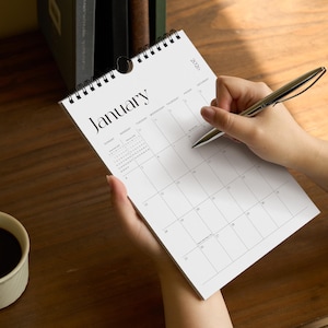 May include: A white wall calendar for January 2020 with black text and a black spiral binding. The calendar features a grid layout for the month with days of the week labeled at the top. A silver pen is positioned over the calendar page.