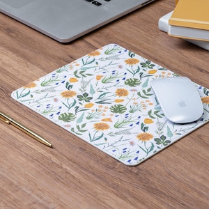 May include: White mousepad with a floral pattern featuring yellow dandelions, bluebells, and green leaves. A white wireless mouse sits on the pad, next to a gold pen. A laptop and a yellow notebook are in the background.