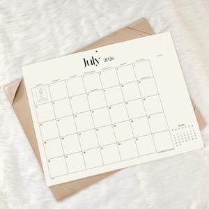 May include: A cream-colored desk calendar for July 2026 featuring a grid layout with days of the week, a small August 2026 calendar in the corner, and a notes section. The calendar is held within a tan-colored protective desk pad cover.