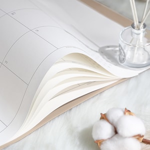 May include: A beige-bound planner with white pages featuring a grid calendar layout. The pages are slightly curled, revealing the internal structure. A clear glass reed diffuser with white sticks and a cotton boll are positioned near the planner.