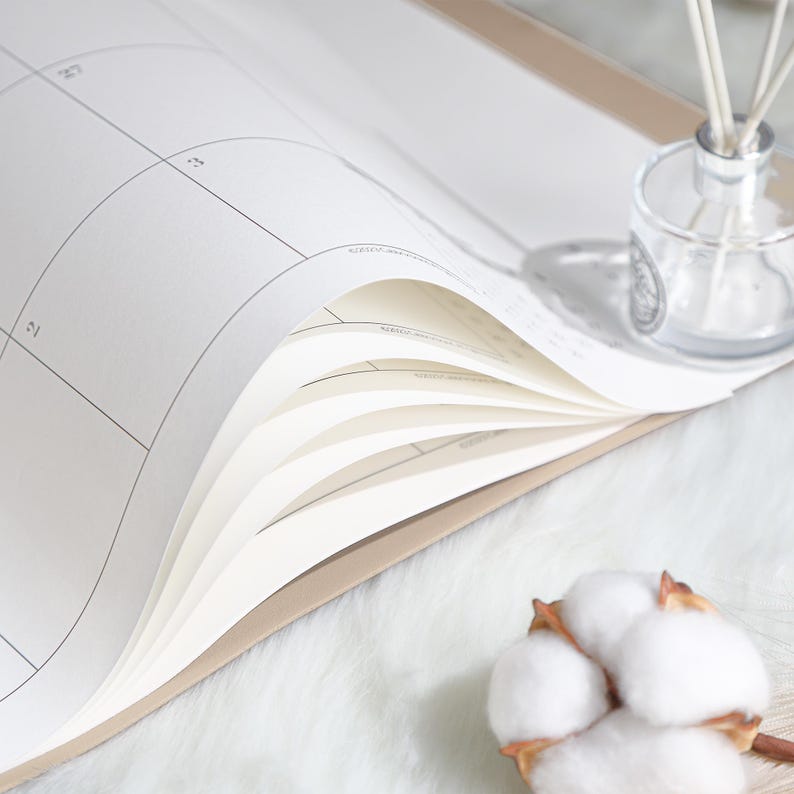 May include: A beige-bound planner with white pages featuring a grid calendar layout. The pages are slightly curled, revealing the internal structure. A clear glass reed diffuser with white sticks and a cotton boll are positioned near the planner.