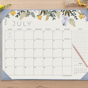 K&ouml;nnte beinhalten: Ein Schreibtischkalender f&uuml;r Juli 2026 mit einer floralen Illustration aus gelben und violetten Blumen am oberen Rand. Der Kalender hat ein Rasterlayout mit einem Notizbereich auf der rechten Seite und hellblauen Eckschonern.