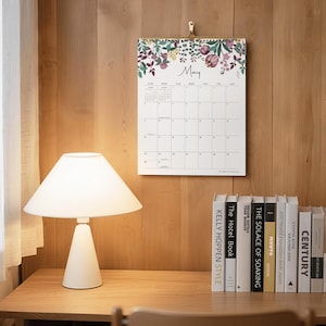 May include: A wall calendar for May 2027 featuring a white background with a floral illustration at the top. The calendar displays a grid for the month with the text May and 2027 clearly visible. It is hung by a gold-colored metal clip.