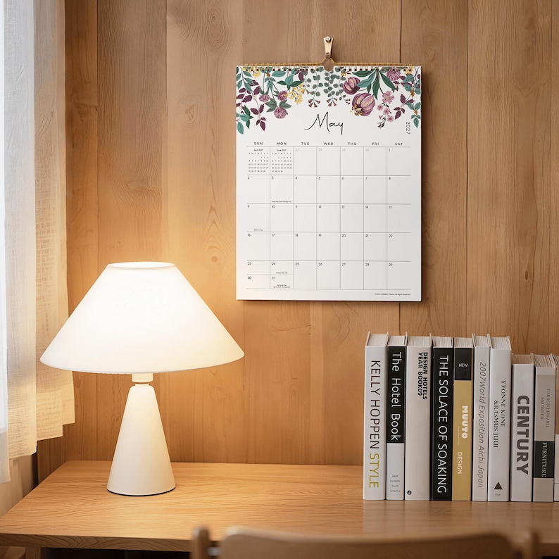 May include: A wall calendar for May 2027 featuring a white background with a floral illustration at the top. The calendar displays a grid for the month with the text May and 2027 clearly visible. It is hung by a gold-colored metal clip.