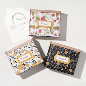 May include: Three rectangular metal recipe tins with wooden lids, each featuring a floral pattern and a gold-toned label holder with the word Recipes. One tin has strawberries, one has yellow flowers on white, and one has flowers on black. Includes a recipe card.
