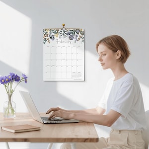 May include: A wall calendar for the month of February featuring a botanical illustration of purple and yellow flowers at the top. The calendar grid displays the days of the week and dates in black text on a white background, hanging from a gold pin.