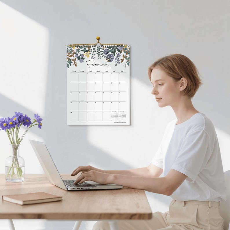 May include: A wall calendar for the month of February featuring a botanical illustration of purple and yellow flowers at the top. The calendar grid displays the days of the week and dates in black text on a white background, hanging from a gold pin.