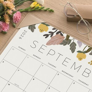K&ouml;nnte beinhalten: Ein Kalenderblatt f&uuml;r September 2026 mit einer Blumenillustration aus gelben Bl&uuml;ten und gedeckten gr&uuml;nen Bl&auml;ttern. Das Kalenderraster zeigt die Wochentage und Daten. Eine Brille mit d&uuml;nnem Rahmen liegt daneben auf dem Untergrund.