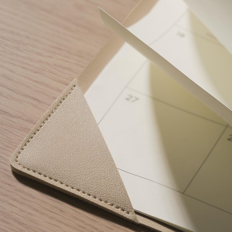 K&ouml;nnte beinhalten: Eine beige, texturierte Ecktasche, die eine cremefarbene Kalenderseite mit sichtbaren Rasterlinien und der in Schwarz gedruckten Zahl 27 h&auml;lt. Die Ecktasche weist entlang der Kanten sichtbare N&auml;hte auf, die das Papierblatt fixieren.