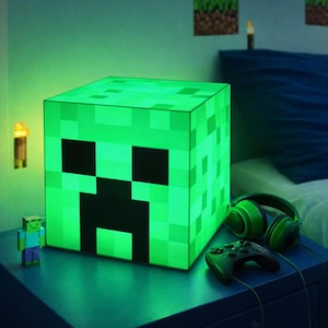 May include: A green pixelated cube lamp with a Creeper face design. The lamp is illuminated, casting a green glow. A Minecraft character figurine, green headphones, and a game controller are on a blue surface.