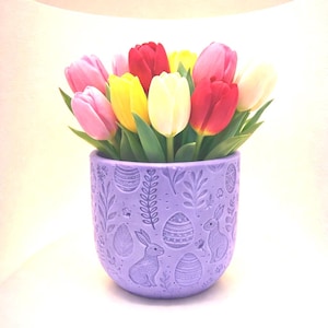 May include: A bouquet of colorful tulips in a lavender ceramic pot. The pot is decorated with embossed Easter-themed designs, including eggs and rabbits. The tulips are a mix of pink, red, yellow, and white.