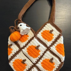 Op de afbeelding: Een gehaakte tas gemaakt van vierkante motieven met oranje pompoenen op een witte achtergrond, omlijst door bruine randen. De tas heeft een bruin handvat en twee kleine bungelende gehaakte bedeltjes: een oranje pompoen en een wit spookje.