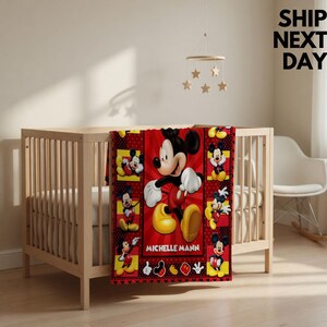 May include: A red and black patterned blanket featuring multiple illustrations of Mickey Mouse in various poses, with the name MICHELLE MANN printed in white capital letters across the centre of the design on the front of a wooden cot.