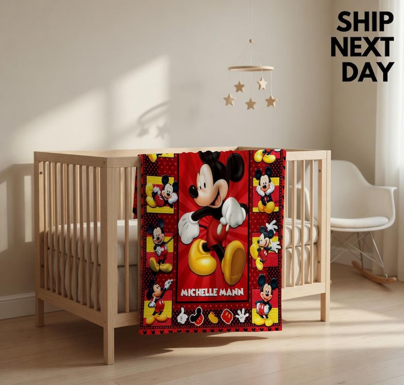 May include: A red and black patterned blanket featuring multiple illustrations of Mickey Mouse in various poses, with the name MICHELLE MANN printed in white capital letters across the centre of the design on the front of a wooden cot.