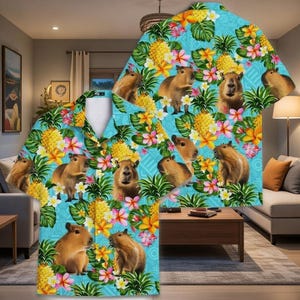 Könnte beinhalten: Ein kurzärmeliges Hemd mit tropischem Muster. Das Hemd hat einen hellblauen Hintergrund mit Capybaras, Ananas und bunten Blumen. Das Muster enthält Gelb-, Rosa-, Grün- und Brauntöne. Das Hemd ist im Button-Down-Stil gehalten.