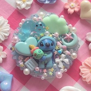 Puede incluir: Una pieza de arte de resina azul con un personaje de dibujos animados sosteniendo una muñeca, rodeada de corazones, nubes, estrellas y perlas de colores pastel. La pieza está sobre un fondo a cuadros rosa y blanco, con flores decorativas.