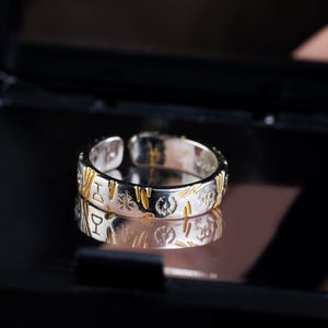 May include: Silver-toned adjustable ring with gold-colored accents and engraved symbols. The ring is displayed in a black box, reflecting its design. The symbols include a wine glass, a star, and other decorative elements.