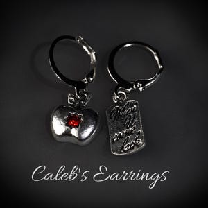 以下が含まれることがあります： レバーバッククロージャー付きのシルバーカラーのイヤリング。片方のイヤリングには赤い宝石が付いたリンゴのチャーム、もう片方にはテキストが入った長方形のチャームが付いています。「Where it came back」の文字が見えます。「Caleb's Earrings」が下に書かれています。