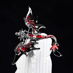 May include: A silver dragon brooch with red and black detailing. The dragon features outstretched wings and red gemstones. The brooch is set against a black backdrop, emphasising its intricate design.