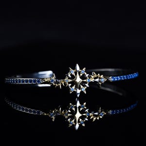 May include: A silver and gold-toned bracelet with a starburst design. The bracelet features a central star motif and is adorned with blue gemstones. The design is mirrored in the reflection, set against a black background.
