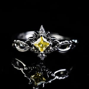 May include: A silver ring with a yellow square-cut gemstone at the center, surrounded by smaller white gemstones and intricate metalwork. The ring's design includes a crown-like top and a reflective surface.