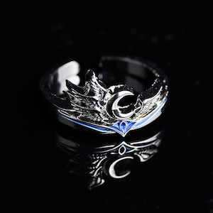 Könnte beinhalten: Ein silberfarbener Ring mit einem himmlischen Design. Der Ring hat Halbmond- und Flügelakzente mit einem blauen Emailleband. Der Ring ist vor einem schwarzen Hintergrund platziert, der seine komplizierten Details hervorhebt.