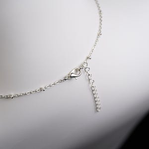 May include: Close-up of a delicate silver necklace with a lobster clasp and an adjustable chain. The necklace features small, round beads along the chain and a heart-shaped extension. The jewelry is displayed against a white background.