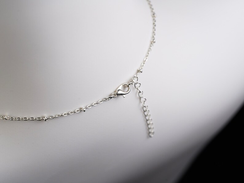 May include: Close-up of a delicate silver necklace with a lobster clasp and an adjustable chain. The necklace features small, round beads along the chain and a heart-shaped extension. The jewelry is displayed against a white background.