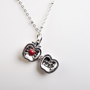 May include: A silver-toned apple-shaped pendant necklace with a red heart-shaped stone inside. The apple opens to reveal a textured interior. The necklace chain is delicate and beaded, creating a charming accessory.