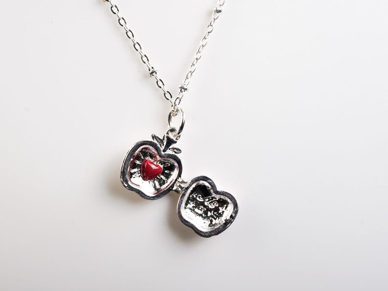 May include: A silver-toned apple-shaped pendant necklace with a red heart-shaped stone inside. The apple opens to reveal a textured interior. The necklace chain is delicate and beaded, creating a charming accessory.